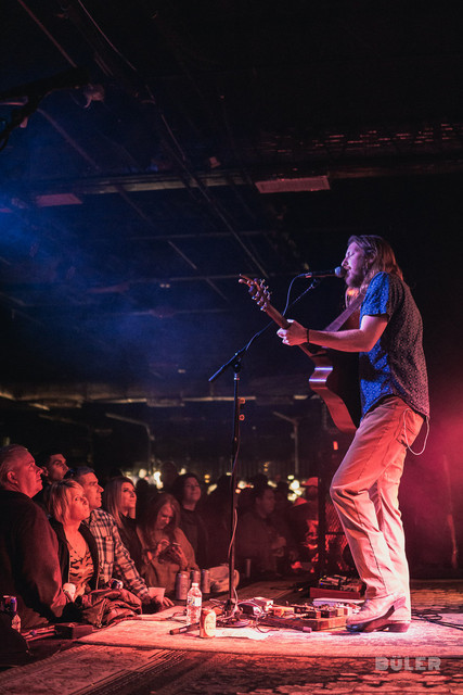 Kenny Freeman singing into a microphone and playing acoustic guitar on a club stage, lit in blue and red.